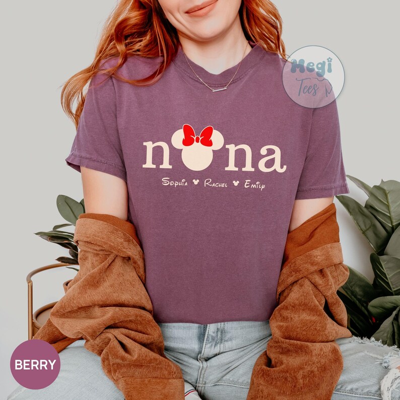 May include: Berry-colored t-shirt with the word "nona" in a white font, featuring a Minnie Mouse head with a red bow. Below the word "nona" are the names Soputa, Rachel, and Emily. The shirt is a short-sleeved crew neck.