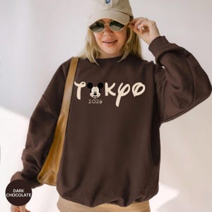 May include: Dark chocolate-colored sweatshirt with the word "Tokyo" in a stylized font, featuring a Mickey Mouse graphic and the year "2020". The sweatshirt is a long-sleeved crewneck.