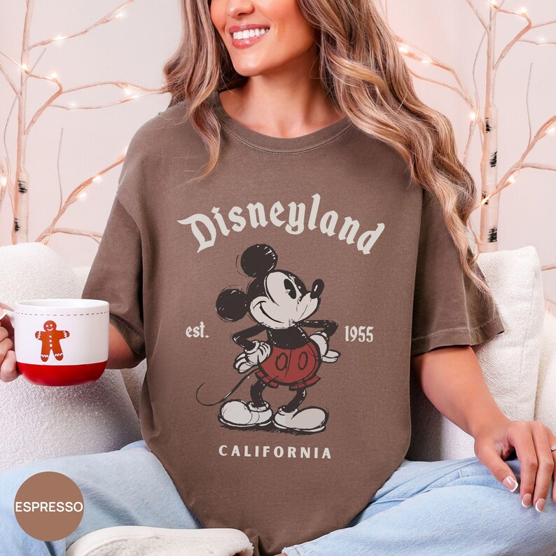 Puede incluir: Camiseta color espresso con un gr&aacute;fico vintage de Disneyland con Mickey Mouse. El dise&ntilde;o incluye el texto "Disneyland", "est. 1955" y "California". La camiseta se combina con una taza blanca y roja con un dise&ntilde;o de hombre de jengibre.