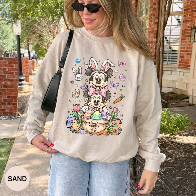 Puede incluir: Una sudadera de color arena presenta un gr&aacute;fico colorido con tem&aacute;tica de Pascua. El dise&ntilde;o incluye los personajes de Mickey y Minnie Mouse con orejas de conejo, rodeados de huevos de Pascua, zanahorias y flores. La palabra "SAND" est&aacute; impresa en la parte inferior.