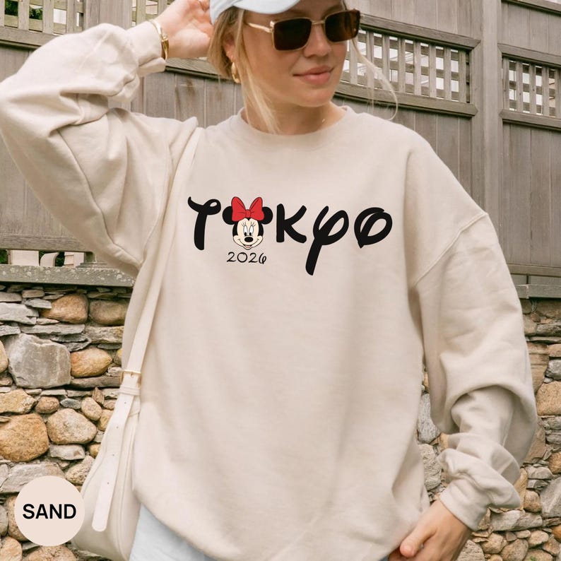 May include: Sand-colored sweatshirt with the word "Tokyo" in black script, featuring a Minnie Mouse graphic with a red bow and the year "2026". The sweatshirt has long sleeves and a crew neck.