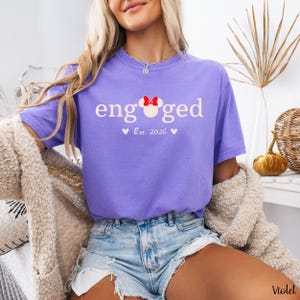 K&ouml;nnte beinhalten: Ein violettes T-Shirt mit dem Wort "engaged" in Wei&szlig;. Eine rote Minnie-Mouse-Schleife befindet sich &uuml;ber dem "g" in "engaged". Darunter steht "Est. 2026" mit zwei kleinen Mickey-Mouse-K&ouml;pfen. Das Shirt wird mit Jeansshorts getragen.