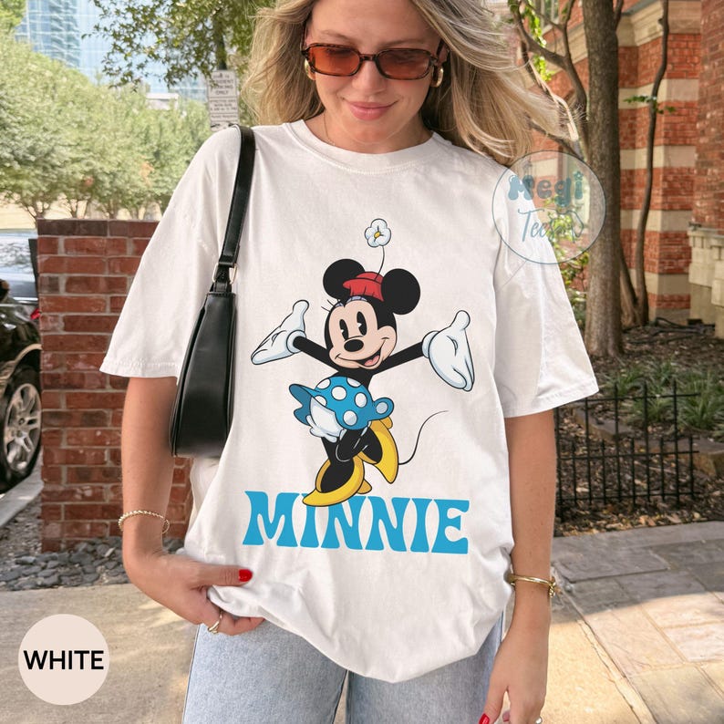 May include: White t-shirt featuring a cartoon Minnie Mouse design. Minnie is depicted with a red hat, blue and white polka dot skirt, and yellow shoes. The word "MINNIE" is printed in blue below the character. The shirt also has the word "WHITE" printed on it.