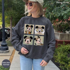 May include: Dark gray heather sweatshirt featuring four Mickey Mouse images in safari attire. The images are set against a light green and beige striped background. The sweatshirt has the text "Mickey Mouse" and "DARK GRAY HEATHER".