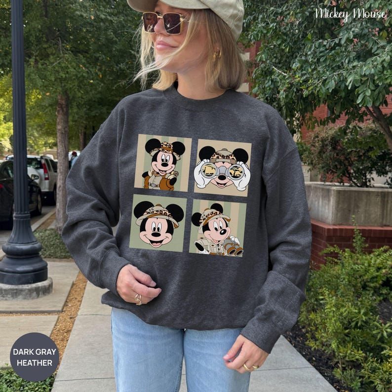 May include: Dark gray heather sweatshirt featuring four Mickey Mouse images in safari attire. The images are set against a light green and beige striped background. The sweatshirt has the text "Mickey Mouse" and "DARK GRAY HEATHER".