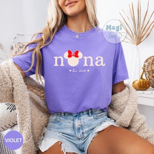 May include: A violet t-shirt with the word "nona" in white, featuring a Minnie Mouse head with a red bow. Below the word is "Est. 2026" with hearts. The shirt is part of the "Hegi Tees" collection.