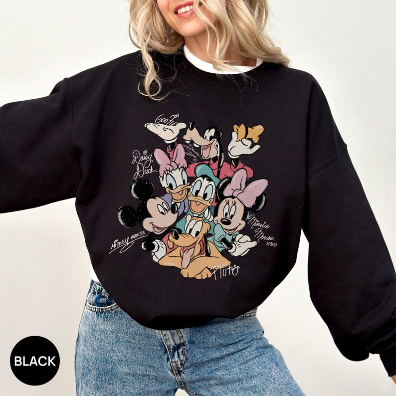 K&ouml;nnte beinhalten: Schwarzes Sweatshirt mit einem farbenfrohen Aufdruck von Disney-Figuren, darunter Mickey Mouse, Minnie Mouse, Donald Duck, Daisy Duck, Goofy und Pluto. Das Sweatshirt hat einen Rundhalsausschnitt und lange &Auml;rmel. Das Wort "BLACK" ist unten links aufgedruckt.