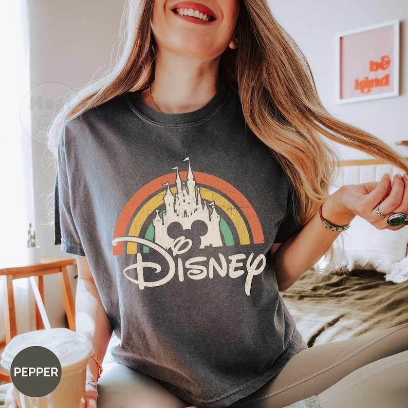 May include: Dark gray t-shirt featuring a colorful rainbow design with a castle, Mickey Mouse silhouette, and the word "Disney" in a vintage style. The shirt has a crew neck and short sleeves.