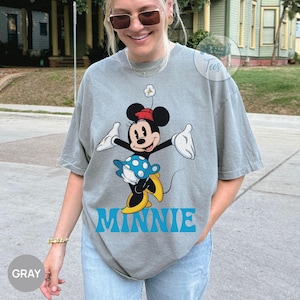 May include: Gray t-shirt featuring a cartoon Minnie Mouse design. Minnie is depicted in a classic pose, wearing a red hat, a blue and white polka-dot skirt, and yellow shoes. The word "MINNIE" is printed in blue below the character.
