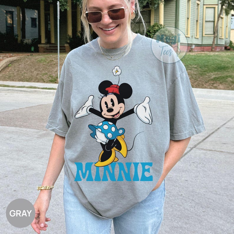 May include: Gray t-shirt featuring a cartoon Minnie Mouse design. Minnie is depicted in a classic pose, wearing a red hat, a blue and white polka-dot skirt, and yellow shoes. The word "MINNIE" is printed in blue below the character.