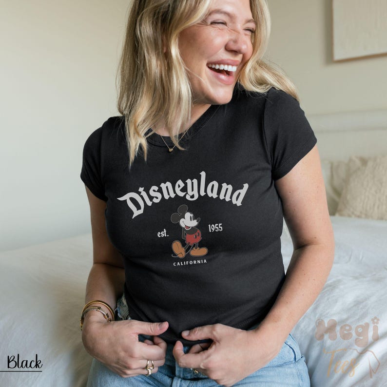 May include: Black t-shirt with the word "Disneyland" in white script, the image of Mickey Mouse, and the text "est. 1955 CALIFORNIA". The shirt has a V-neck and short sleeves.