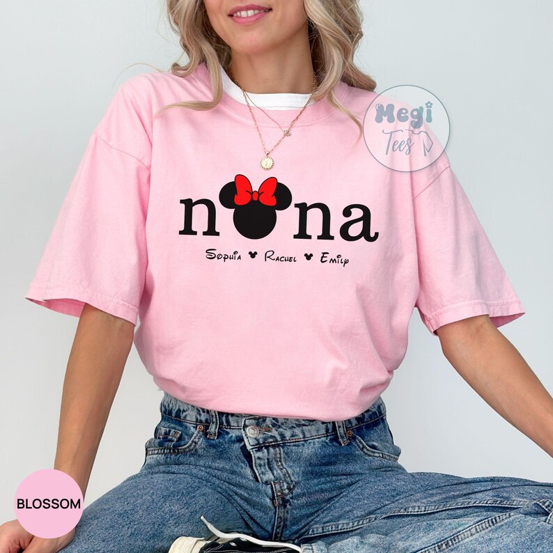 May include: A light pink t-shirt with the word "nona" in black letters, a Minnie Mouse head with a red bow, and three names below. The shirt has a white collar and is made of a soft material.