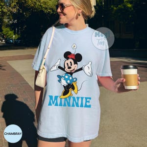 May include: Light blue t-shirt featuring a graphic of Minnie Mouse in a playful pose. The design includes Minnie's signature bow, dress, and yellow shoes, with the name "MINNIE" in blue block letters. The shirt has a relaxed fit.