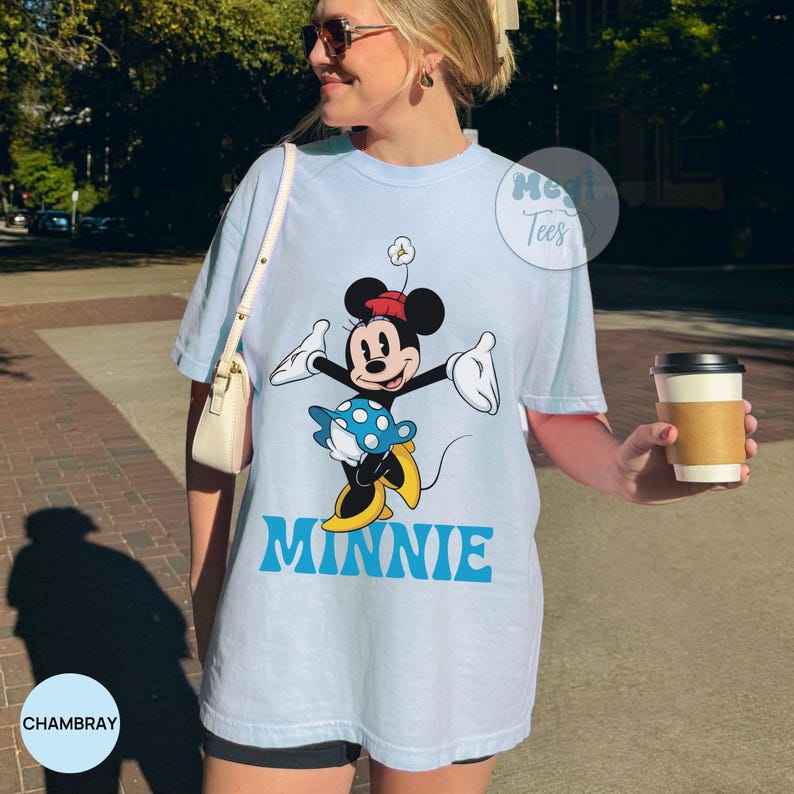 May include: Light blue t-shirt featuring a graphic of Minnie Mouse in a playful pose. The design includes Minnie's signature bow, dress, and yellow shoes, with the name "MINNIE" in blue block letters. The shirt has a relaxed fit.