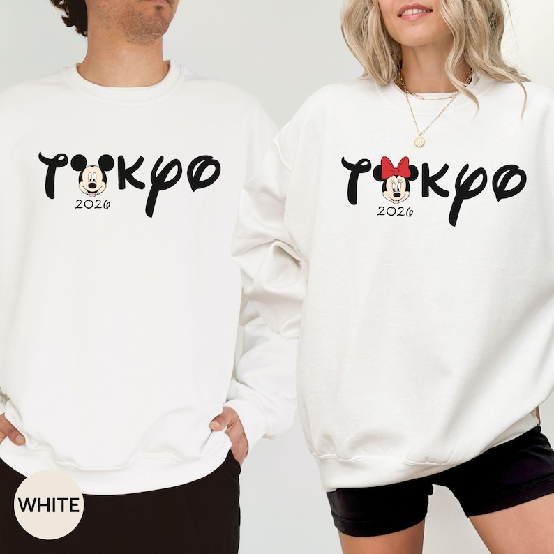 May include: White sweatshirts with the word "Tokyo" in a stylized font, featuring Mickey and Minnie Mouse designs. The year "2026" is printed below. One sweatshirt has a Mickey Mouse design, the other has a Minnie Mouse design with a red bow.
