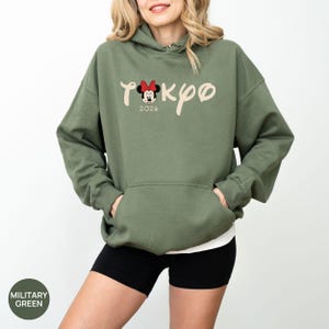 May include: Military green hoodie with a kangaroo pocket. The front features the word "Tokyo" in a script font, with a Minnie Mouse graphic and the year "2020". The hoodie is worn with black shorts.