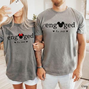 K&ouml;nnte beinhalten: Zwei graue T-Shirts mit dem Wort "engaged" in Schwarz aufgedruckt, wobei ein Mickey Mouse-Kopf das "o" ersetzt. Die Shirts haben auch "Est. 2026" darunter aufgedruckt. Ein Shirt hat eine rote Schleife.