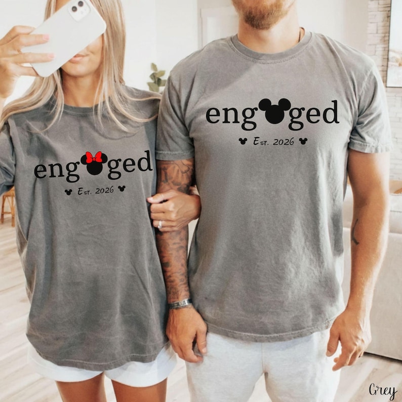 K&ouml;nnte beinhalten: Zwei graue T-Shirts mit dem Wort "engaged" in Schwarz aufgedruckt, wobei ein Mickey Mouse-Kopf das "o" ersetzt. Die Shirts haben auch "Est. 2026" darunter aufgedruckt. Ein Shirt hat eine rote Schleife.