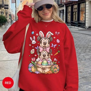 Puede incluir: Una sudadera roja con un dise&ntilde;o de Mickey y Minnie Mouse disfrazados de conejitos de Pascua. El dise&ntilde;o incluye huevos de Pascua, zanahorias y flores. La palabra "RED" est&aacute; impresa en la parte inferior izquierda.
