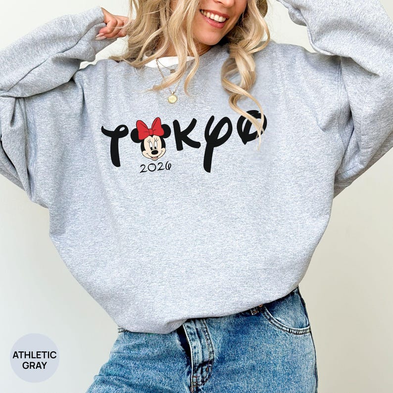 May include: Athletic gray sweatshirt with the word "Tokyo" in black script, featuring a Minnie Mouse graphic with a red bow and the year "2026". The sweatshirt is a light gray color.