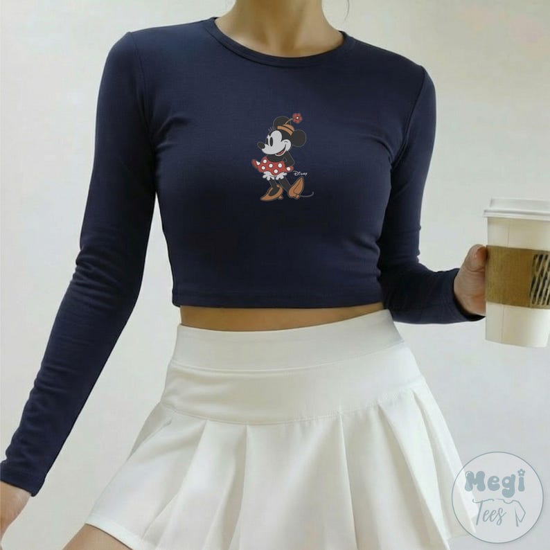 Puede incluir: Top corto de manga larga azul marino con un gr&aacute;fico de Minnie Mouse. El gr&aacute;fico muestra a Minnie Mouse con un vestido de lunares rojos y blancos y un sombrero a juego. El top se combina con una falda plisada blanca. Tambi&eacute;n se ve una taza de caf&eacute;.