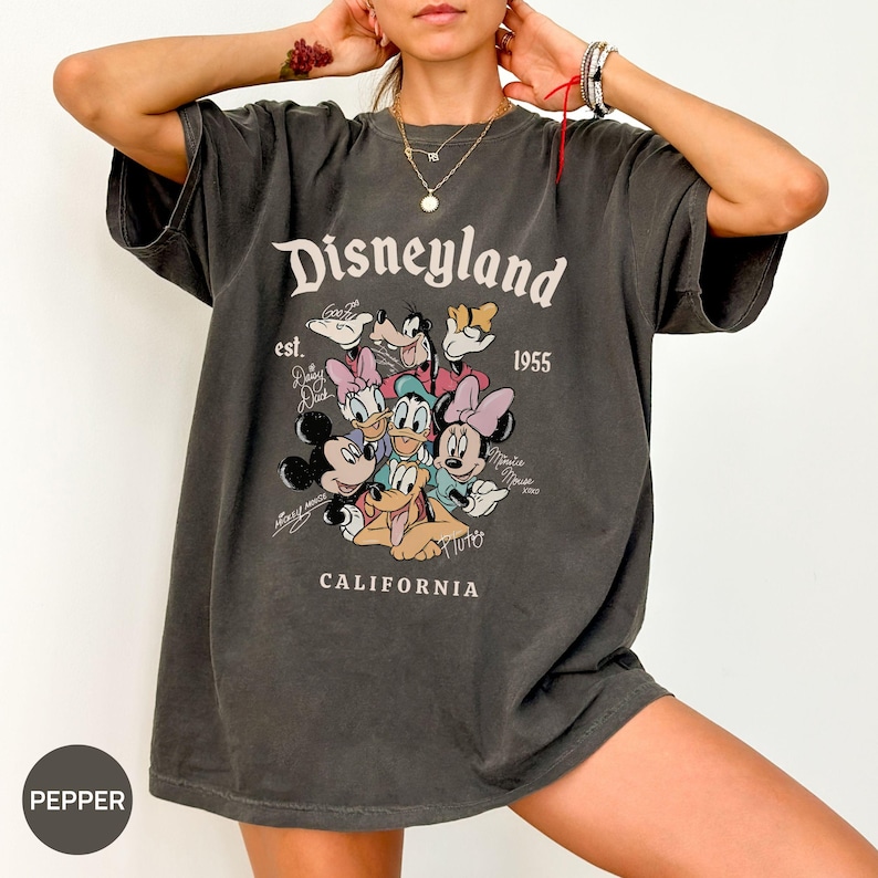 Puede incluir: Camiseta gris oscuro con el logotipo de Disneyland, el texto "est. 1955" y un gr&aacute;fico de personajes de Disney. La palabra "CALIFORNIA" est&aacute; impresa debajo del gr&aacute;fico. La camiseta tiene mangas cortas.