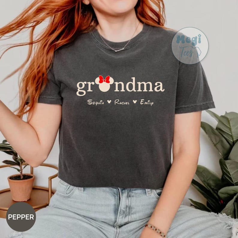 May include: Dark gray t-shirt with the word "grandma" in white script, featuring a red bow graphic. Below the word are three names: Sophia, Rachel, and Emily. The shirt is a casual style.