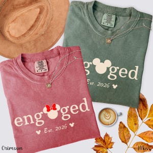 K&ouml;nnte beinhalten: Zwei Comfort Colors T-Shirts, eines in Karmesinrot und eines in Moosgr&uuml;n, mit dem Aufdruck "engaged". Das karmesinrote Shirt hat eine Minnie Mouse Schleife. Beide Shirts haben "Est. 2026" und einen Mickey Mouse Kopf. Ein brauner Hut, ein Latte und Herbstbl&auml;tter sind ebenfalls im Bild.