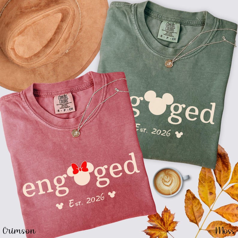 K&ouml;nnte beinhalten: Zwei Comfort Colors T-Shirts, eines in Karmesinrot und eines in Moosgr&uuml;n, mit dem Aufdruck "engaged". Das karmesinrote Shirt hat eine Minnie Mouse Schleife. Beide Shirts haben "Est. 2026" und einen Mickey Mouse Kopf. Ein brauner Hut, ein Latte und Herbstbl&auml;tter sind ebenfalls im Bild.