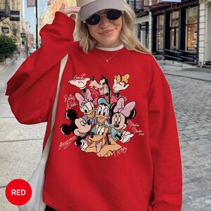 K&ouml;nnte beinhalten: Ein rotes Sweatshirt mit einer Grafik von Disney-Figuren, darunter Micky Maus, Minnie Maus, Donald Duck, Daisy Duck, Goofy und Pluto. Das Wort "RED" befindet sich in einem roten Kreis unten links.