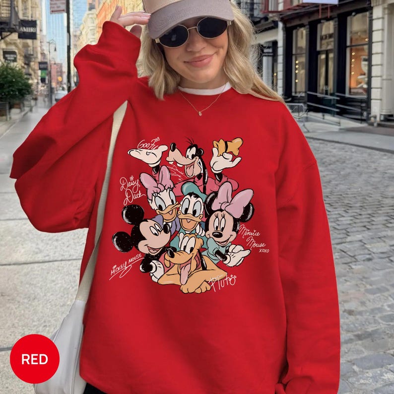 K&ouml;nnte beinhalten: Ein rotes Sweatshirt mit einer Grafik von Disney-Figuren, darunter Micky Maus, Minnie Maus, Donald Duck, Daisy Duck, Goofy und Pluto. Das Wort "RED" befindet sich in einem roten Kreis unten links.
