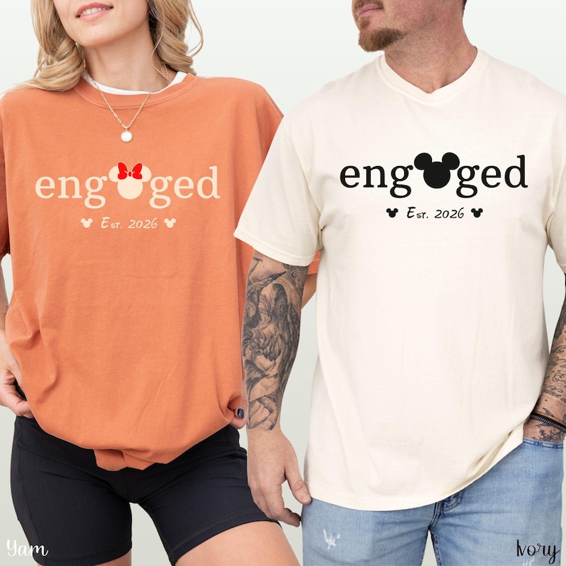 K&ouml;nnte beinhalten: Zwei T-Shirts, eines orangefarbenes und eines elfenbeinfarbenes, mit dem Wort "engaged" bedruckt. Das Wort "engaged" ist in einer verspielten Schriftart mit einem Mickey Mouse-Kopf, der das "o" ersetzt. Darunter steht "Est. 2026" mit Herzen.