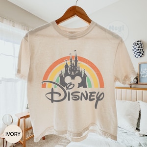 May include: Ivory-colored t-shirt featuring a colorful rainbow design with a castle silhouette and the word "Disney" in a script font. The shirt is hanging on a wooden hanger. The overall theme is Disney.