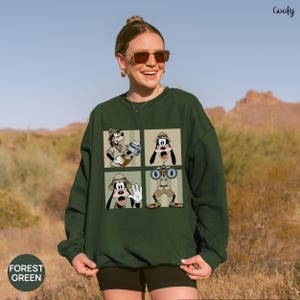 May include: Forest green sweatshirt featuring a four-panel graphic of Goofy in safari attire. Each panel shows Goofy in a different pose, with binoculars, a camera, and a waving gesture. The word "Coofy" is in the top right corner.