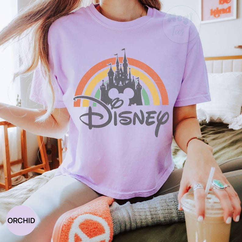 May include: A light purple t-shirt featuring a colorful rainbow design with a castle silhouette and the word "Disney" in a script font. The shirt is a soft, comfortable material.