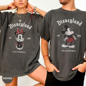 Puede incluir: Dos camisetas gris carb&oacute;n con el logotipo de Disneyland y gr&aacute;ficos de Mickey y Minnie Mouse. Las camisetas tienen el texto "Disneyland", "est. 1955" y "CALIFORNIA" impreso. Las camisetas son usadas por dos personas.