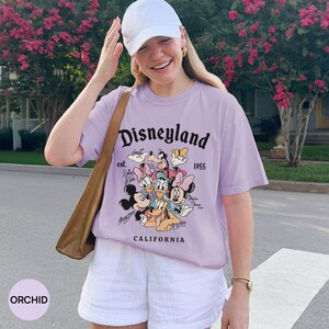 Puede incluir: Camiseta de color orqu&iacute;dea con un gr&aacute;fico de Disneyland que presenta a Mickey Mouse, Minnie Mouse, Donald Duck y otros personajes. La camiseta tiene el texto "Disneyland est. 1955 California". El modelo lleva una gorra y pantalones cortos blancos.