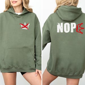 May include: Olive green hoodie with a front pocket and a hood. The front features a red and white graphic of a crown with a line through it. The back of the hoodie displays the word "NOPE" in white with a red crown graphic.