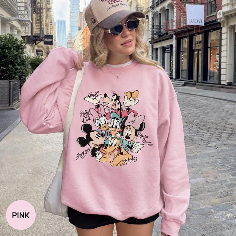 K&ouml;nnte beinhalten: Hellrosa Sweatshirt mit einer Grafik von Disney-Figuren, darunter Micky und Minnie Maus, Donald Duck und Goofy. Das Sweatshirt hat lange &Auml;rmel und einen Rundhalsausschnitt. Das Wort "PINK" befindet sich in einem rosa Kreis unten links.