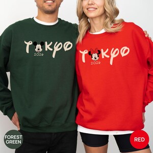May include: Forest green and red sweatshirts with the word "Tokyo" in a cursive font, featuring Mickey and Minnie Mouse. The year "2026" is below the characters. The forest green sweatshirt has Mickey Mouse, and the red sweatshirt has Minnie Mouse.