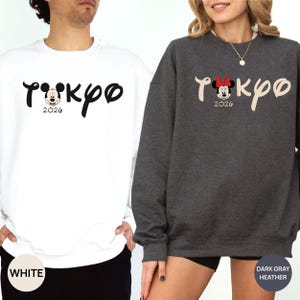 May include: White and dark gray heather sweatshirts with the word "Tokyo" in a script font, featuring a Mickey Mouse graphic and the year "2026". The white sweatshirt has black text, while the dark gray one has beige text and a Minnie Mouse graphic.