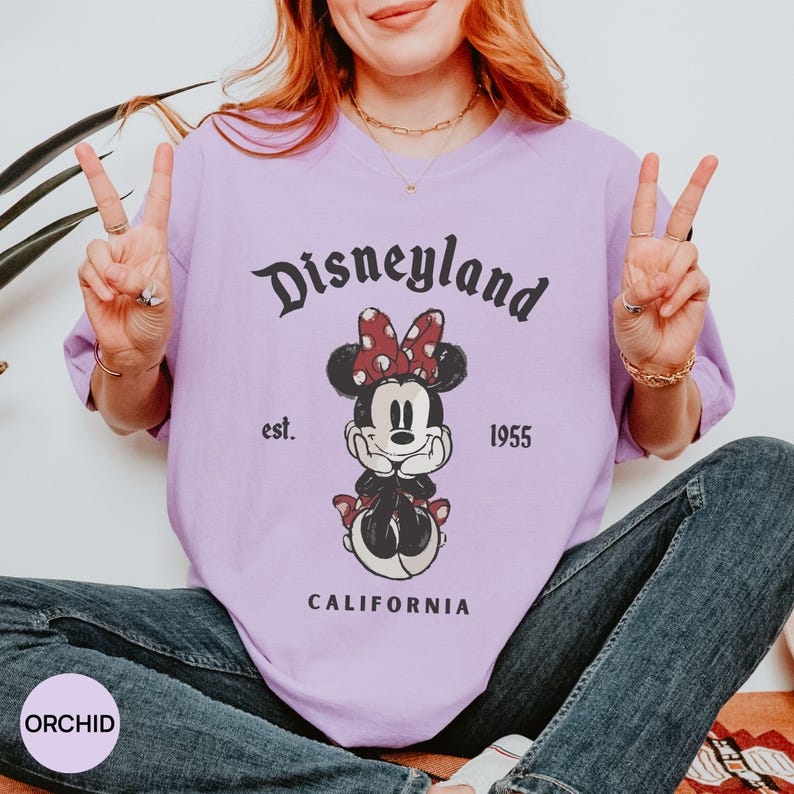 Puede incluir: Una camiseta de color lila claro con un dise&ntilde;o de Minnie Mouse con un lazo rojo de lunares y la palabra "Disneyland" encima. El texto "est. 1955 CALIFORNIA" tambi&eacute;n est&aacute; impreso en la camiseta. La camiseta es de color s&oacute;lido.