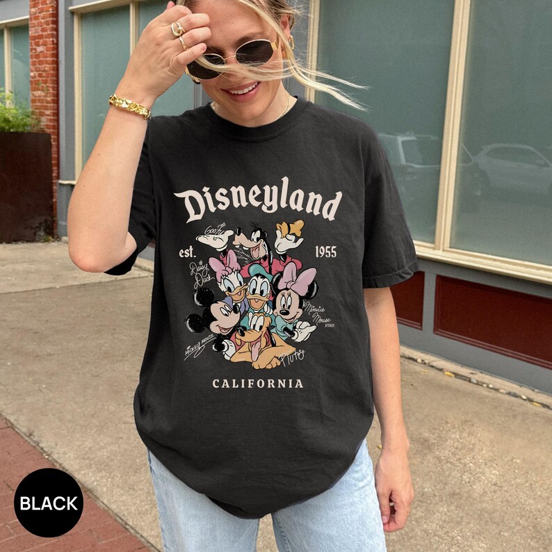 Puede incluir: Camiseta negra con el logo de Disneyland y un gr&aacute;fico de personajes de Disney. La camiseta incluye el texto "est. 1955" y "CALIFORNIA". La camiseta es de cuello redondo y tiene mangas cortas.
