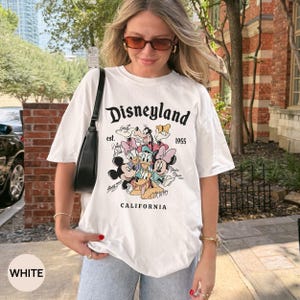 Puede incluir: Camiseta blanca con un gr&aacute;fico de Disneyland que presenta a Mickey Mouse, el Pato Donald y otros personajes de dibujos animados. La camiseta incluye el texto "Disneyland est. 1955 CALIFORNIA".