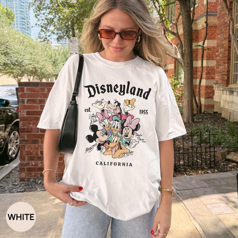 Puede incluir: Camiseta blanca con un gr&aacute;fico de Disneyland que presenta a Mickey Mouse, el Pato Donald y otros personajes de dibujos animados. La camiseta incluye el texto "Disneyland est. 1955 CALIFORNIA".