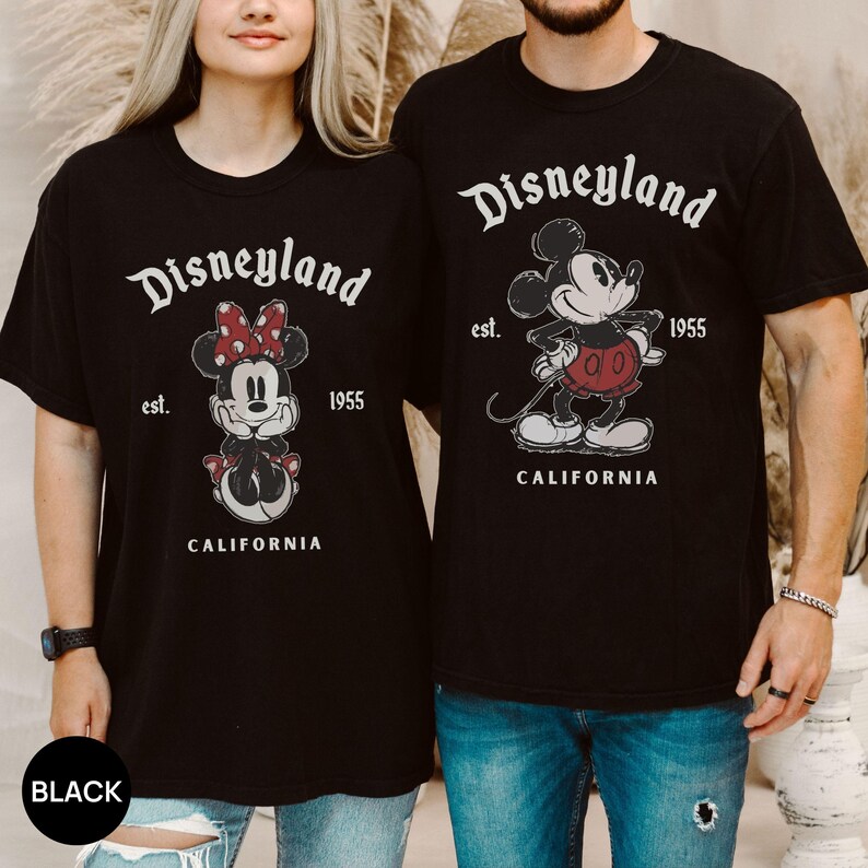 Puede incluir: Camisetas negras con gr&aacute;ficos de Disneyland. Una camiseta muestra a Minnie Mouse, la otra a Mickey Mouse. Ambas camisetas tienen el texto "Disneyland" encima del personaje, con "est. 1955 CALIFORNIA" debajo.