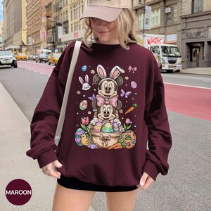 Puede incluir: Sudadera granate con un dise&ntilde;o tem&aacute;tico de Pascua. El dise&ntilde;o presenta a Mickey y Minnie Mouse como conejos, rodeados de huevos de Pascua, zanahorias y flores. La palabra "MAROON" est&aacute; impresa en la parte inferior izquierda.