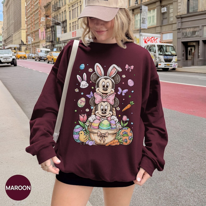 Puede incluir: Sudadera granate con un dise&ntilde;o tem&aacute;tico de Pascua. El dise&ntilde;o presenta a Mickey y Minnie Mouse como conejos, rodeados de huevos de Pascua, zanahorias y flores. La palabra "MAROON" est&aacute; impresa en la parte inferior izquierda.