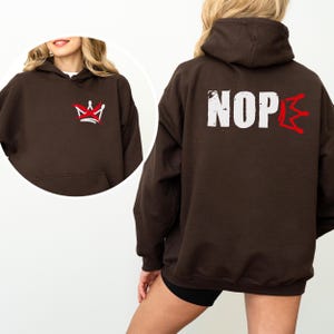 May include: A dark brown hoodie with a front pocket and a hood. The front features a red and white crown design. The back displays the word "NOPE" in white block letters with a red lightning bolt-like design replacing the "E".