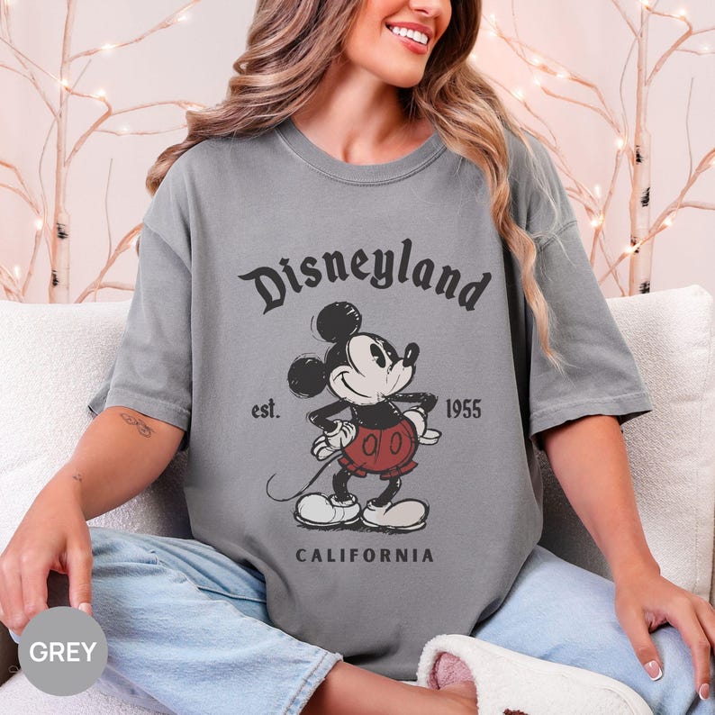 Puede incluir: Camiseta gris con un gr&aacute;fico vintage de Mickey Mouse y el texto "Disneyland" arriba y "California" abajo. La camiseta tambi&eacute;n incluye el texto "est. 1955".